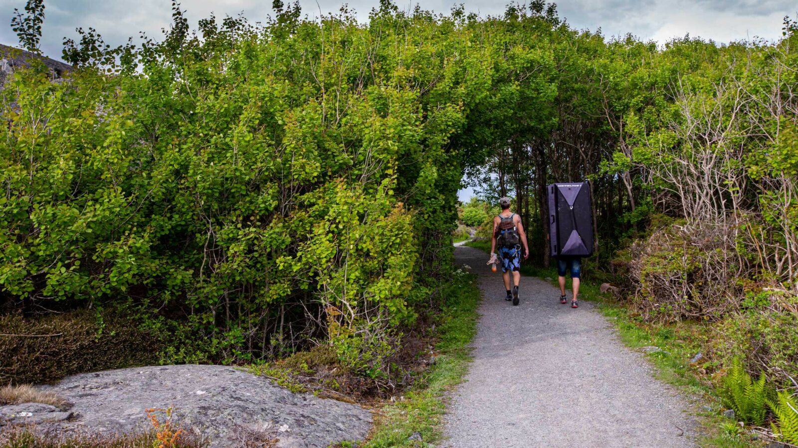 Två klättrare på en stig som passerar genom ett buskage. En av klättrarena bär en crash pad.